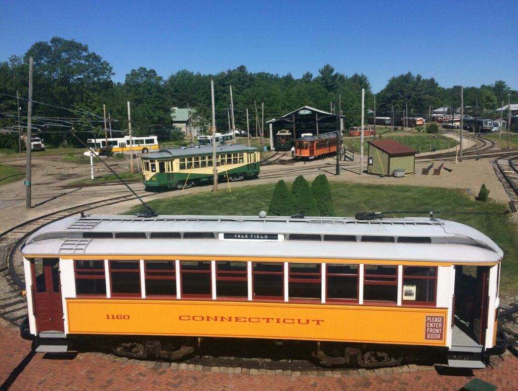 A yellow trolley parked on Seashore Trolley Museum's electric railroad, with the campus and other trolleys in the background.